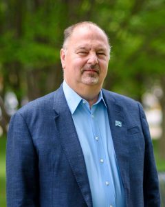 Portrait of Ken Zelazny, Huron Chamber Board Chair. Blue shirt, blue blazer, greeen grass and trees behind.